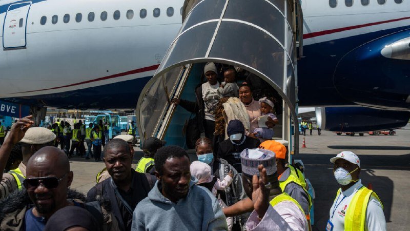 Passengers, including women and children, descend a covered airstair from a parked passenger aircraft onto a tarmac where ground crew in high-visibility vests and masks stand nearby.