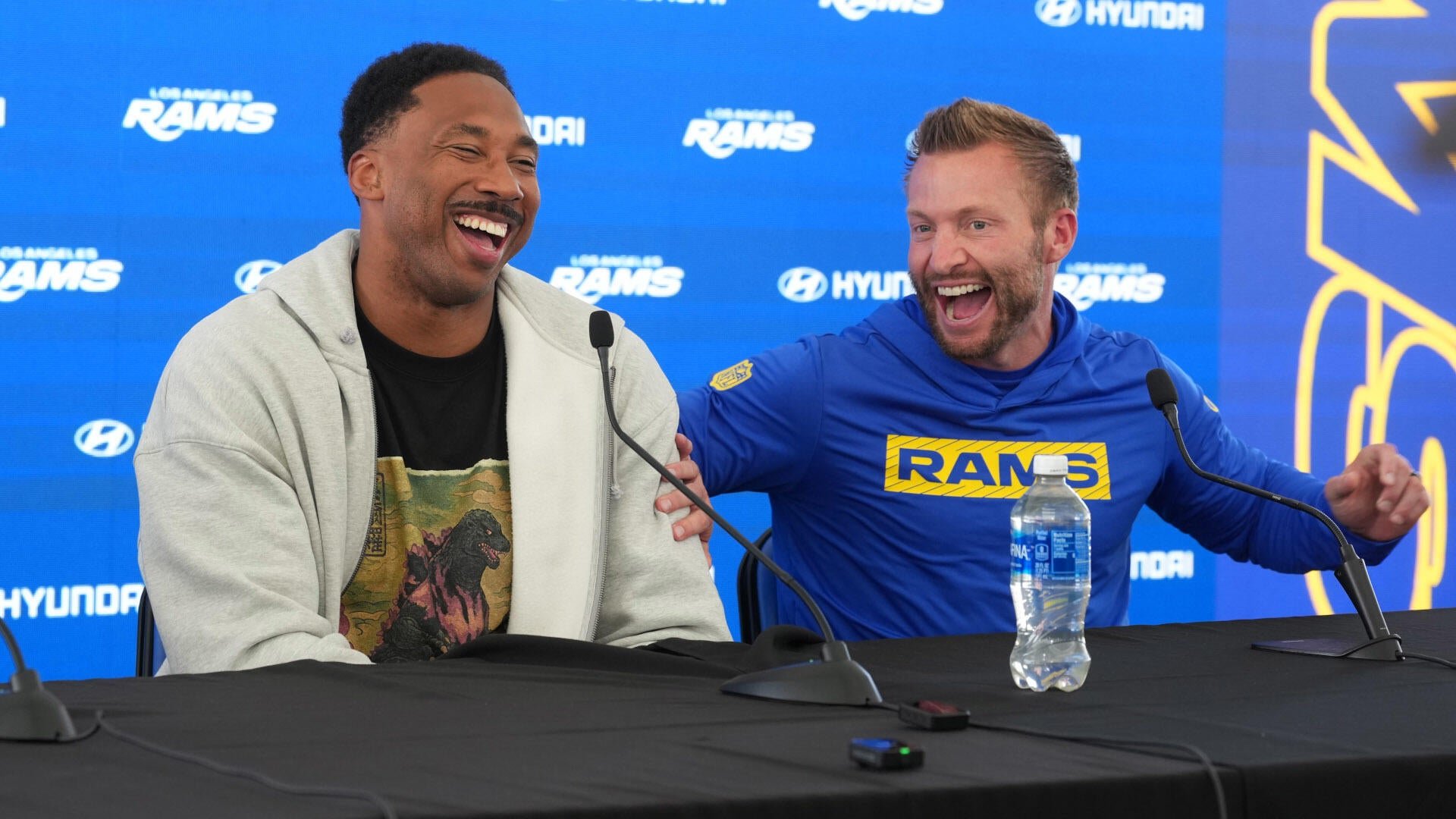 Two men laugh heartily while seated at a press conference table with microphones, against a blue backdrop featuring Los Angeles Rams and Hyundai logos.