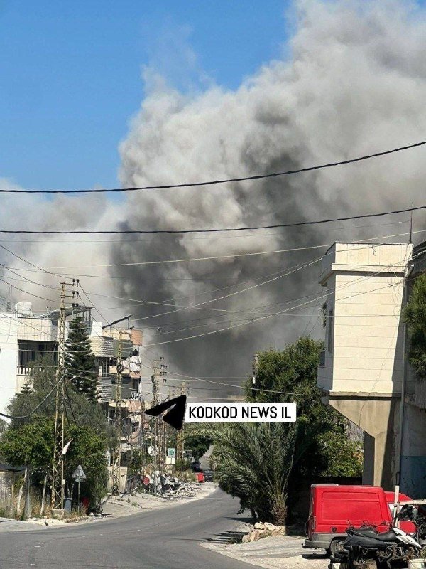 Thick gray smoke billows into the sky above a residential street lined with buildings, power lines, and a parked red van.