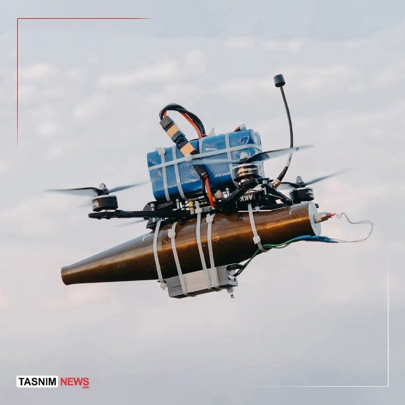A small quadcopter drone carrying a brown cylindrical object secured with straps and blue battery packs hovers mid-air against a cloudy sky.