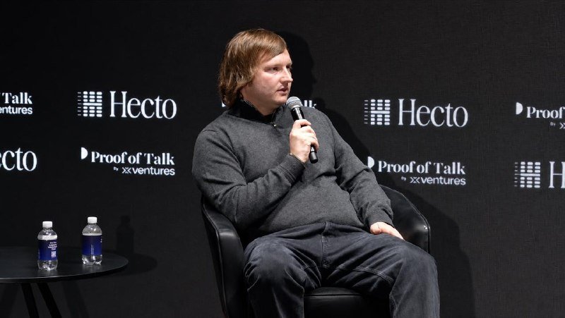 A man in a gray sweater speaks into a microphone while seated on stage, with "Hecto" and "Proof of Talk by xVentures" logos displayed on the backdrop.