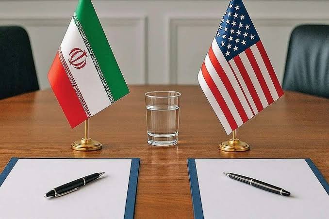 The image is an illustration showing miniature Iranian and American flags on a wooden table, with a glass of water and two blank documents with pens placed between them.