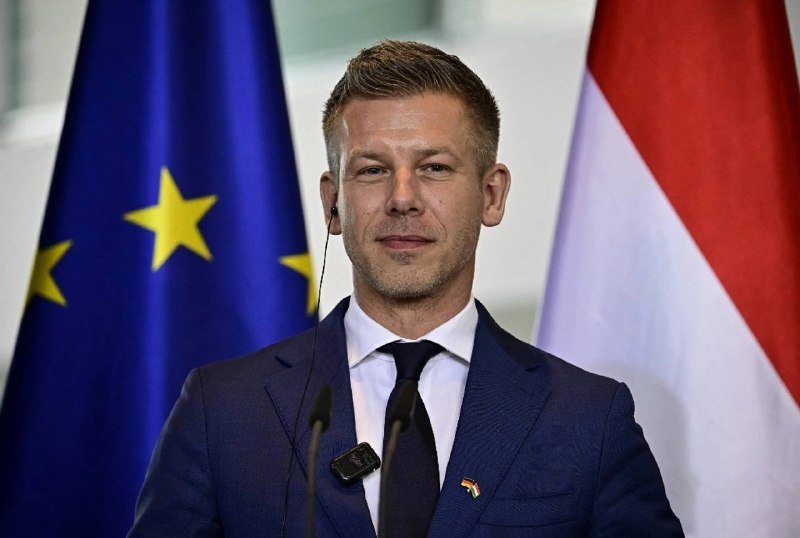 A man in a navy suit with an earpiece stands at a microphone, flanked by an EU flag and a red-white-red flag in the background.