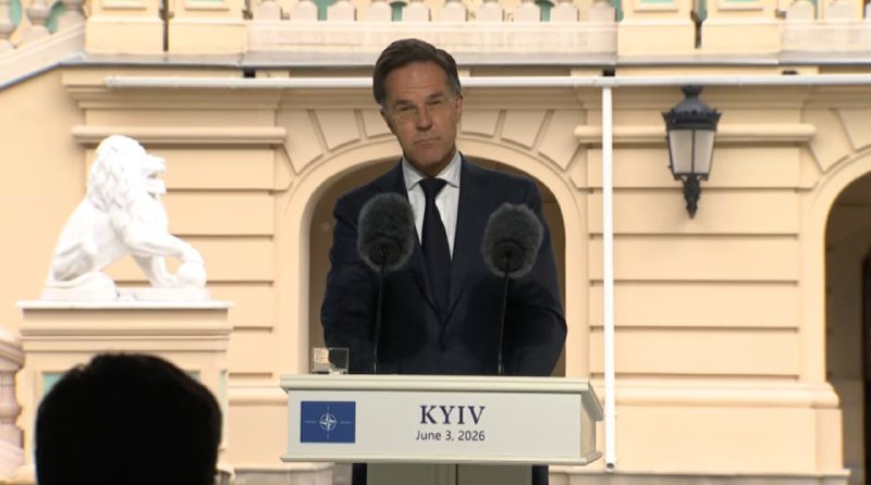 A man in a dark suit and tie stands at a podium labeled "KYIV June 3, 2026" with a NATO logo, speaking into two microphones outside a building with a stone lion statue.