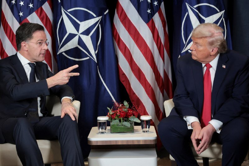 Two men in suits sit facing each other in armchairs, with one gesturing with two fingers, against a backdrop of American and NATO flags.