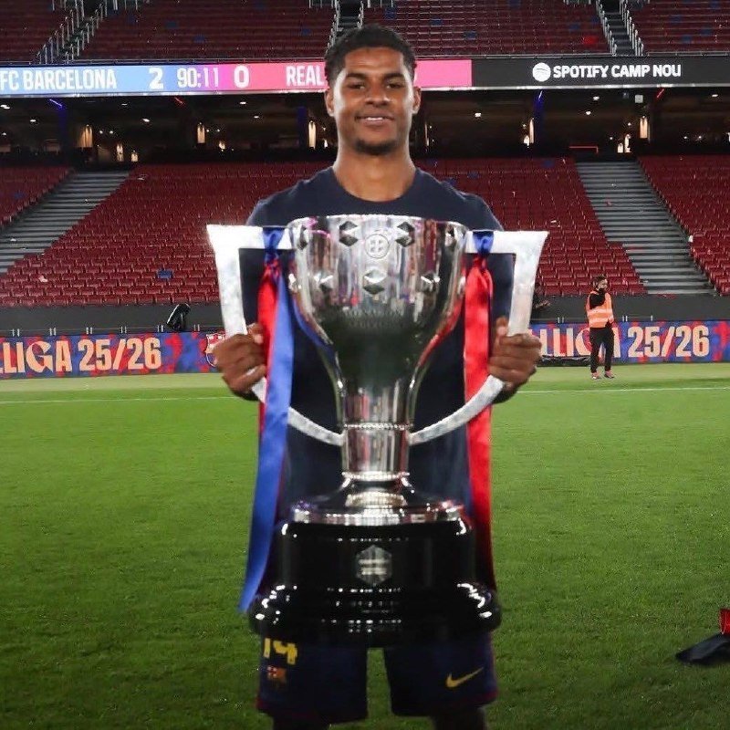 A smiling soccer player in a dark jersey holds a large silver trophy with red and blue ribbons on a stadium field.