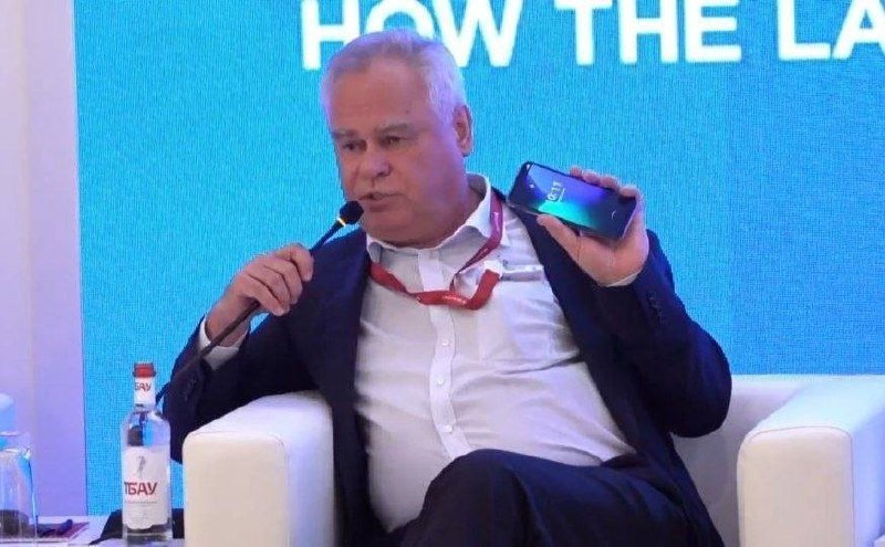 A gray-haired man in a dark suit holds up a smartphone while speaking into a microphone during a panel discussion, with a water bottle and partial text "HOW THE LA" visible behind him.
