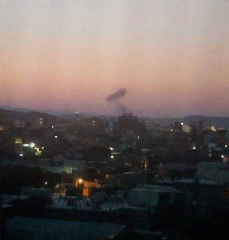 A plume of dark smoke rises above a densely packed city at dusk, with scattered lights visible among the buildings.