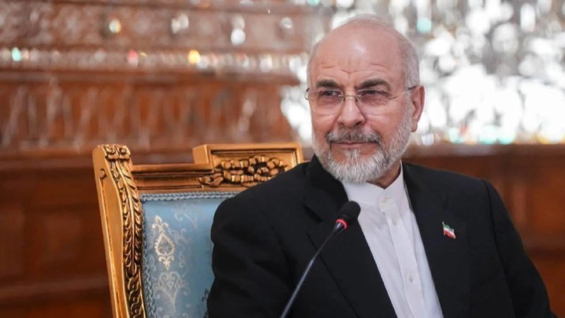 A gray-bearded man in a dark suit sits before a microphone at an ornate blue and gold chair, with a small flag pin on his lapel.