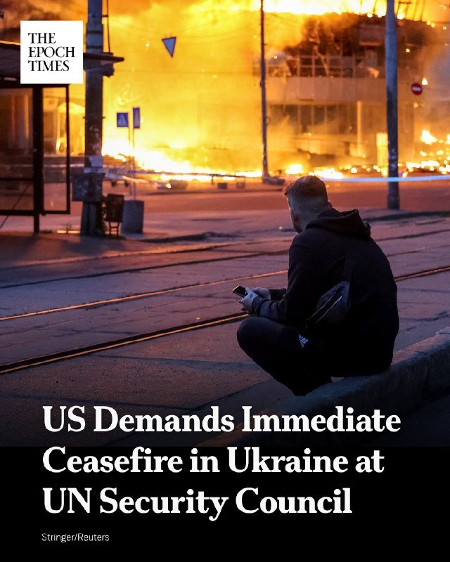 A man sits on a curb looking at his phone as a large fire burns across a street in the background, overlaid with "The Epoch Times" logo and a headline.