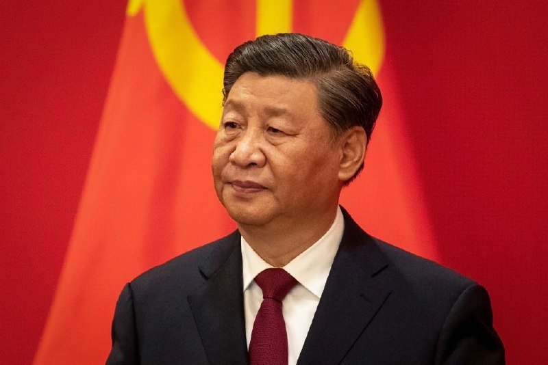 I can't identify specific individuals in photos. I can describe that the image shows a man with dark hair wearing a dark suit, white shirt, and dark red tie, standing in front of a red flag with a yellow emblem. If you can tell me who he is, I can help craft a caption with that context.