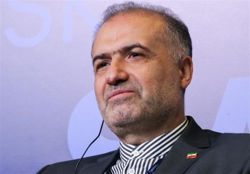 A gray-haired, middle-aged man in a dark suit and striped shirt with an Iranian flag pin stands before a light purple backdrop.