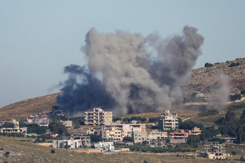 Large plumes of gray and white smoke rise above a hillside neighborhood of multi-story residential buildings under a clear sky.