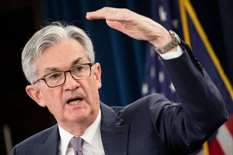 A gray-haired man in a dark suit, glasses, and wristwatch gestures with his raised left hand while speaking, with an American flag visible in the background.
