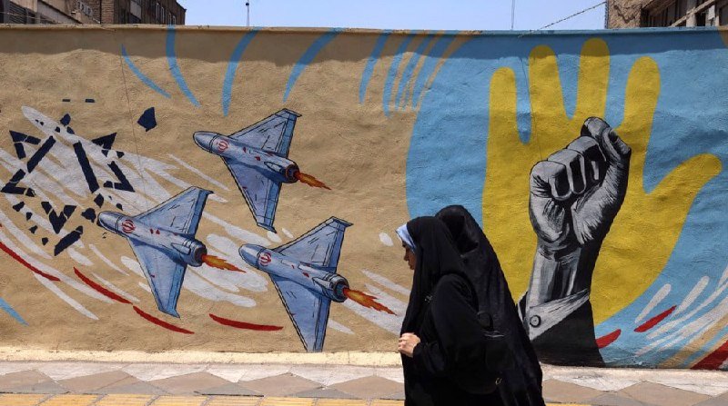 Two women in black chadors walk past a mural depicting fighter jets and a raised fist against a yellow hand shape on an outdoor wall.