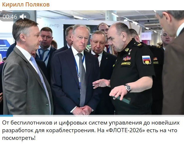 Several men in suits and military uniforms gather at an indoor exhibition, listening to an officer in uniform gesturing while speaking.