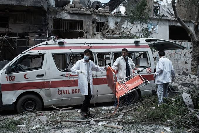 Three men in white coats carry a stretcher beside a damaged "Civil Defense" ambulance marked "102" in front of a heavily destroyed building.