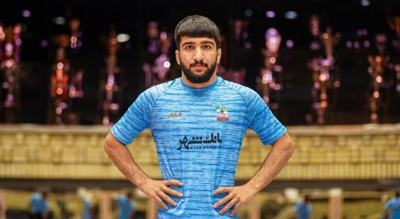 A bearded athlete in a blue jersey stands with hands on hips in an indoor arena, with blurred figures and a wrestling mat visible in the background.