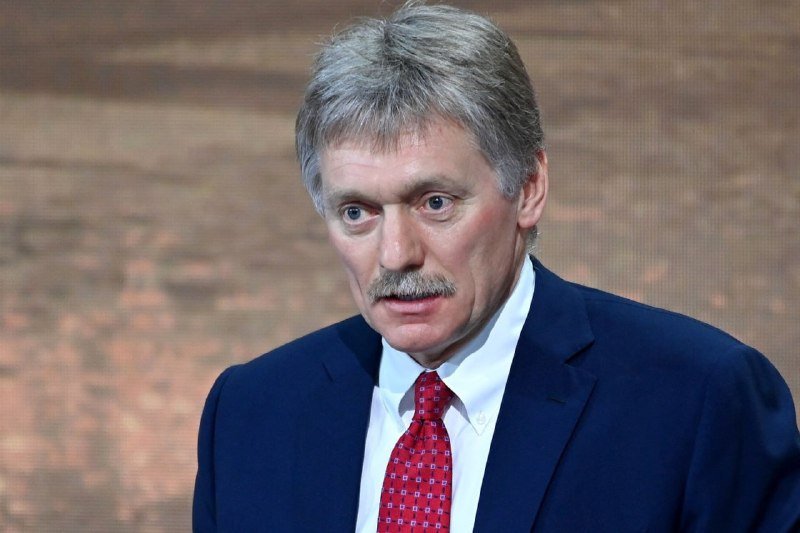 A gray-haired man in a navy suit and red patterned tie speaks against a textured brown background.