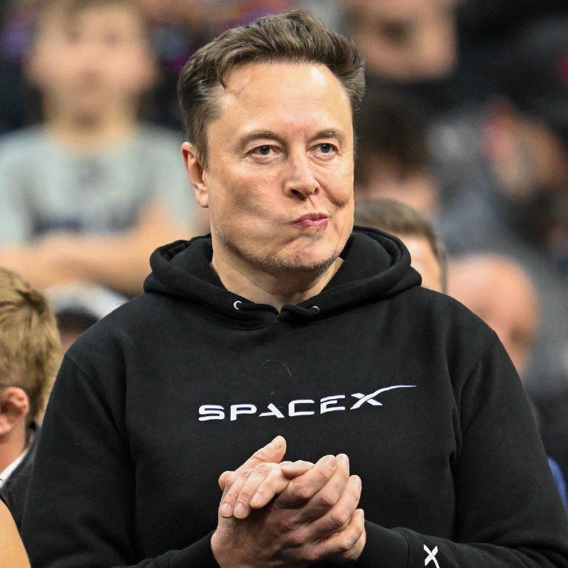 A man wearing a black SpaceX hoodie stands among a crowd, hands clasped together while looking forward.