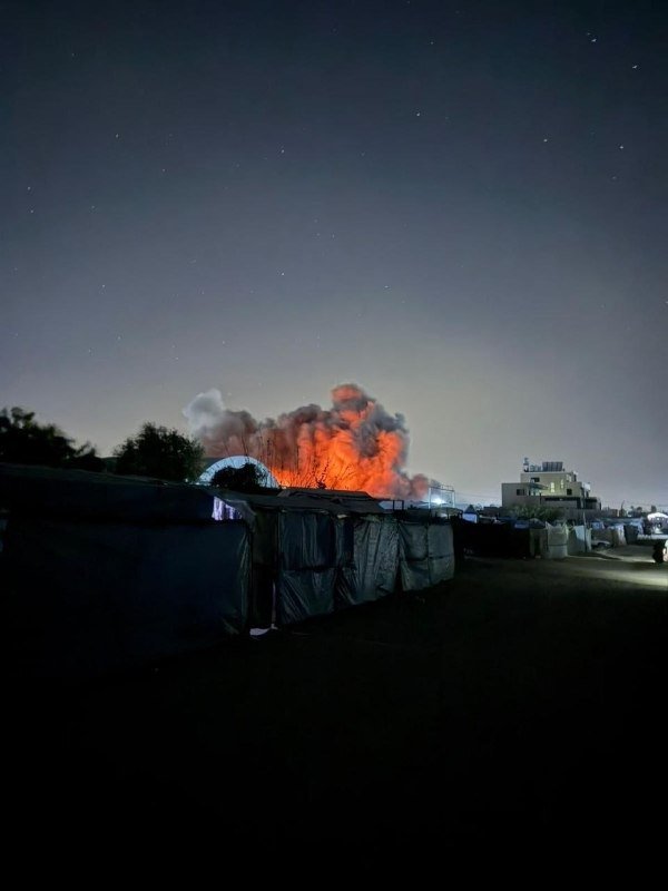 Orange flames and thick smoke rise at night beyond a row of makeshift tents, with illuminated buildings visible in the distance under a dark sky.