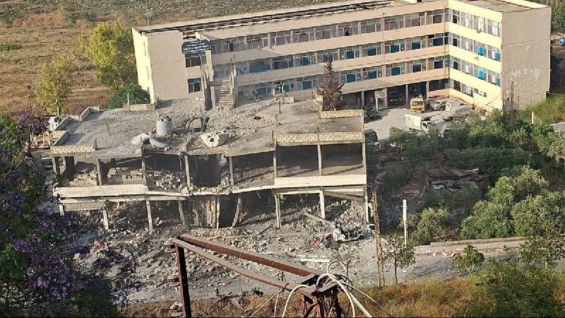 A partially destroyed multi-story building with collapsed walls and rubble sits adjacent to an intact, undamaged multi-story building, with several vehicles parked nearby in a debris-strewn area.