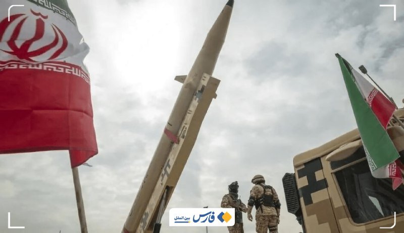 Two soldiers stand beside a camouflaged truck displaying a missile and the Iranian flag against a cloudy sky.