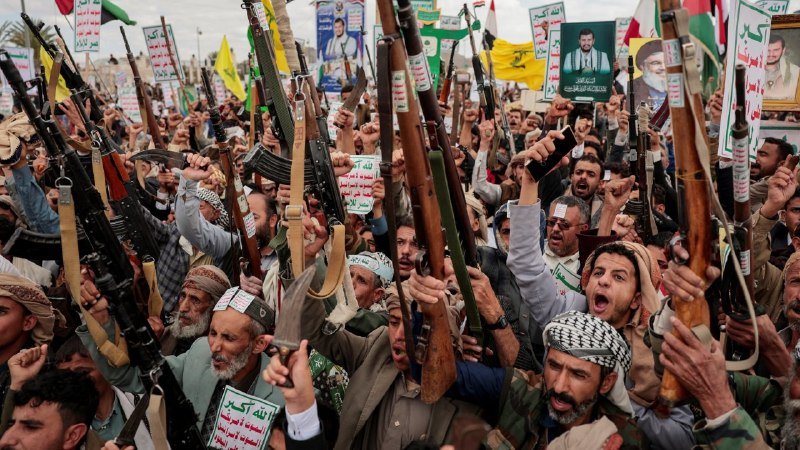 A large crowd of men chant and raise rifles alongside flags and posters bearing portraits and Arabic script during an outdoor demonstration.