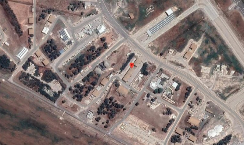 An aerial satellite image shows a large rectangular building complex circled with a dotted white line, surrounded by smaller structures, roads, and trees.