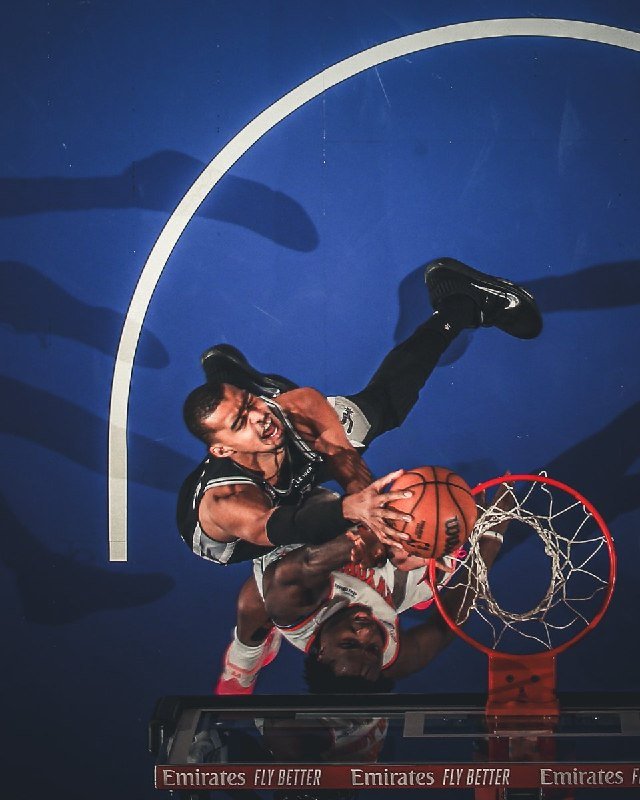 Two basketball players in white and black uniforms compete for the ball mid-air above a red rim, photographed from below against a blue court with "Emirates FLY BETTER" signage visible.
