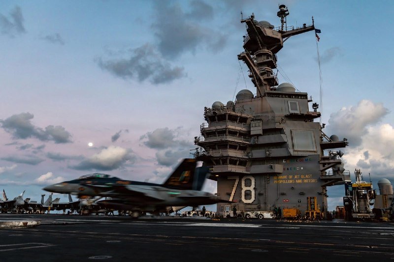 A military jet marked "78" launches off the flight deck of an aircraft carrier at dusk, with other aircraft and crew visible on deck beneath a cloudy sky.