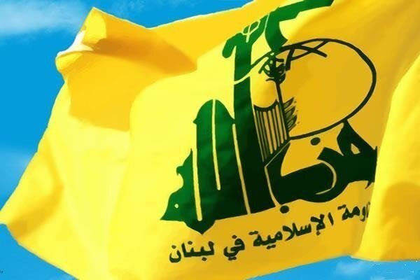 A yellow flag bearing a green emblem with Arabic script and a fist holding a rifle waves against a blue sky.