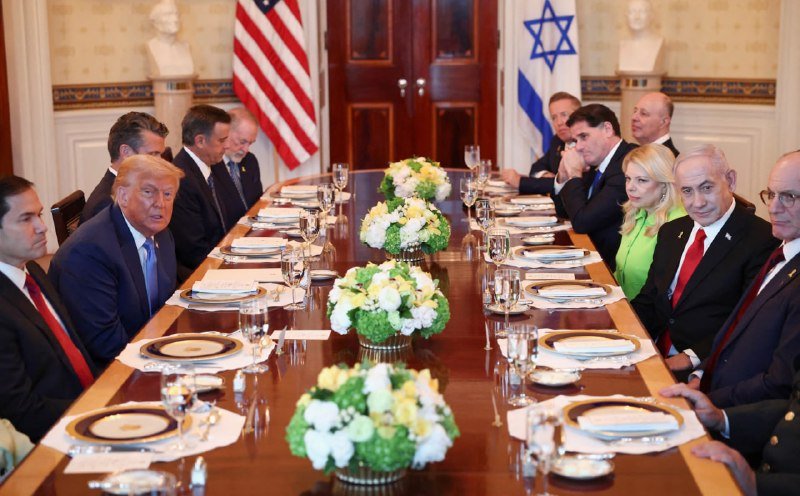 Officials in business attire sit on both sides of a long dining table set with floral centerpieces, with American and Israeli flags displayed in the background.