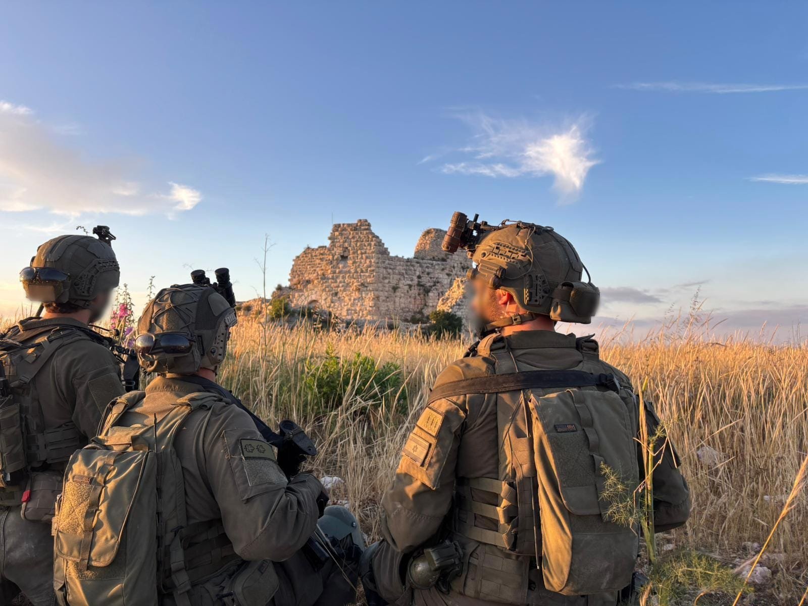 Three soldiers in tactical gear and helmets observe a stone fortress ruin from a grassy field.