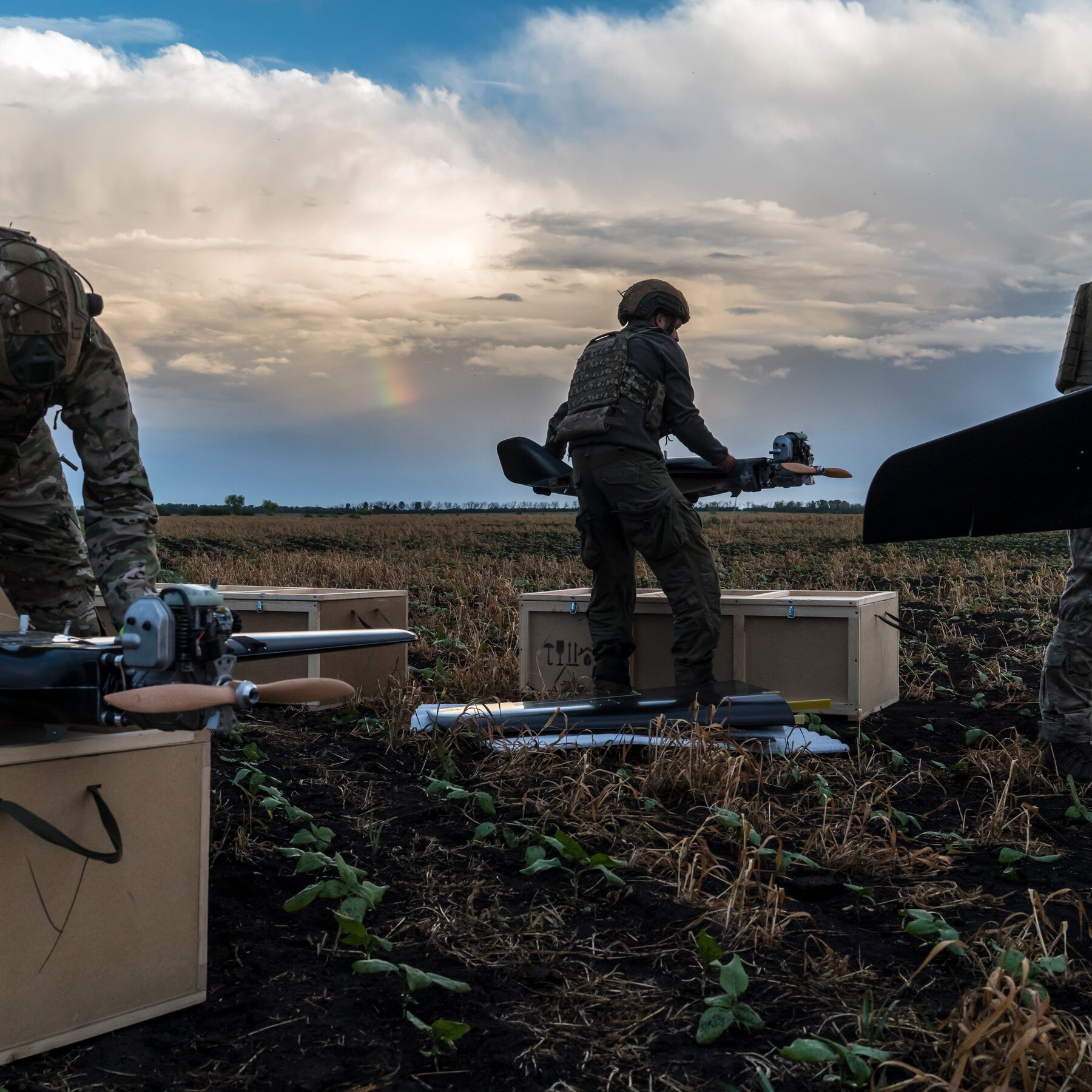 Soldiers in camouflage and tactical gear carry drone-like unmanned aerial vehicles across a harvested field toward wooden crates under a cloudy sky with a faint rainbow.