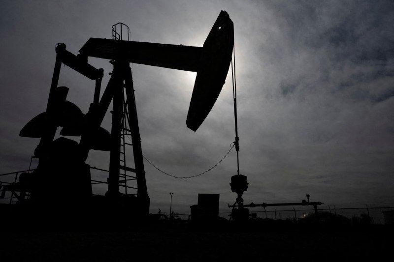 A silhouetted oil pumpjack operates against a cloudy sky with the sun shining behind it.