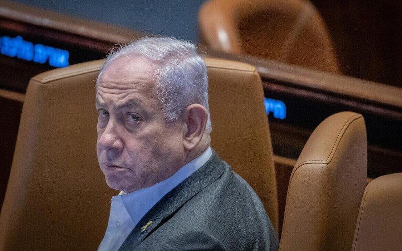 A man in a suit with an Israeli flag pin sits in a brown leather chair, looking sideways.