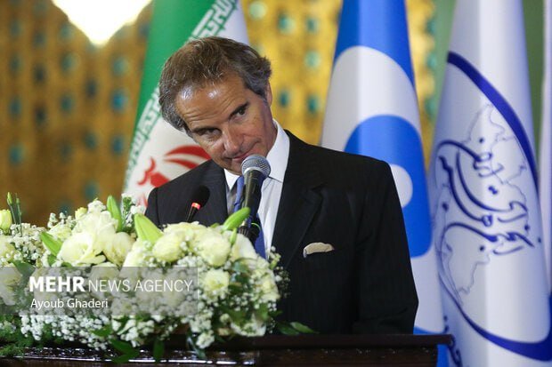 A bald man in a dark suit and tie speaks into a UN-branded microphone against a blue UN emblem backdrop.