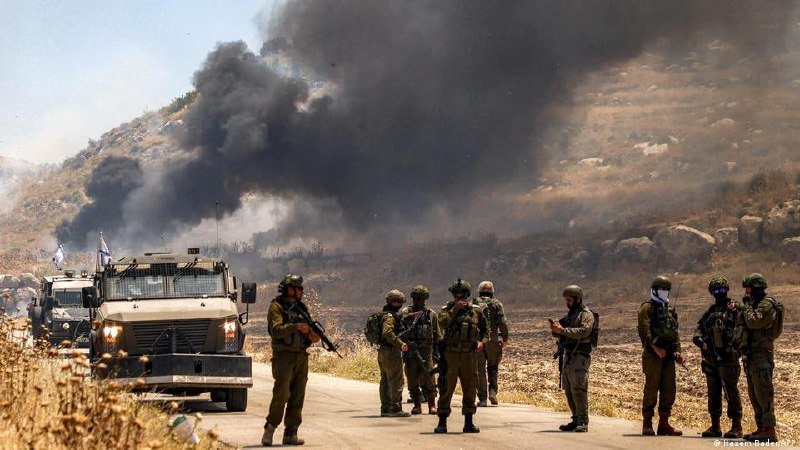 Soldiers in combat gear stand along a road as thick black smoke rises from a hillside behind a military vehicle.