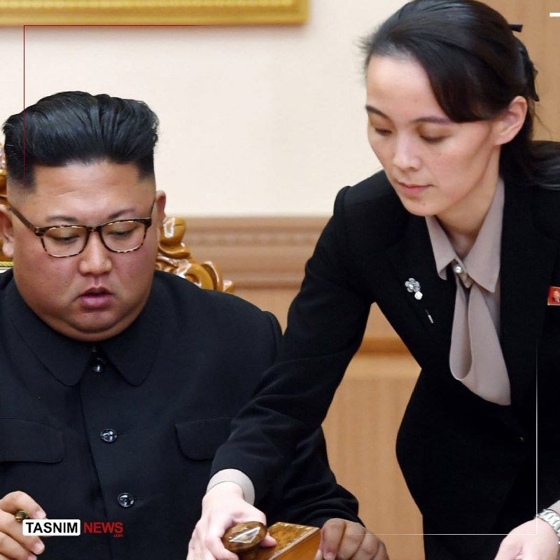 A bespectacled man in a dark suit sits beside a woman in a black blazer who leans over to examine a wooden box on a table, with a "TASNIM NEWS" watermark visible.