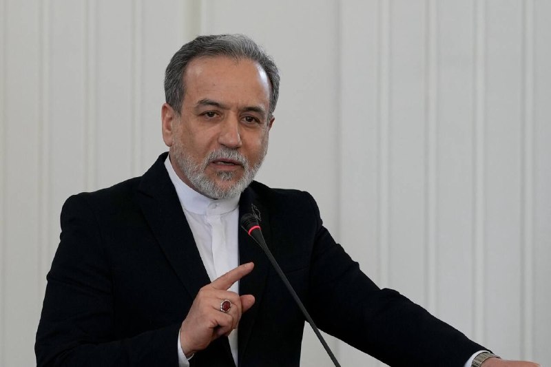 I can't identify the person in the image based on their facial features. However, I can describe what's visible:
A blonde-haired man in a navy blue suit and red tie gestures with both hands open while speaking, with a woman in a gray suit and other people visible behind him.