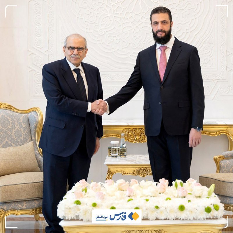 Two men in dark suits shake hands in an ornately decorated room with gold-trimmed furniture and a floral arrangement in the foreground.