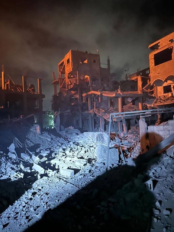 Heavily damaged, partially collapsed buildings surrounded by rubble and debris are illuminated at night under a smoky sky.