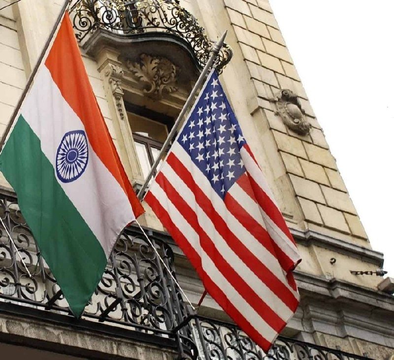 The Indian and American flags hang side by side from an ornate stone building with wrought-iron balconies.