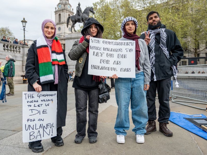 Four people wearing keffiyehs and hijabs stand outdoors holding handwritten signs reading "The government were willing to let us die for an unlawful ban."