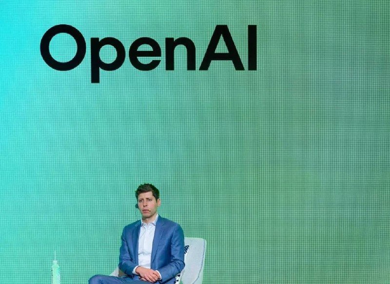 A man in a blue suit sits on a chair in front of a large green screen displaying the "OpenAI" logo.
