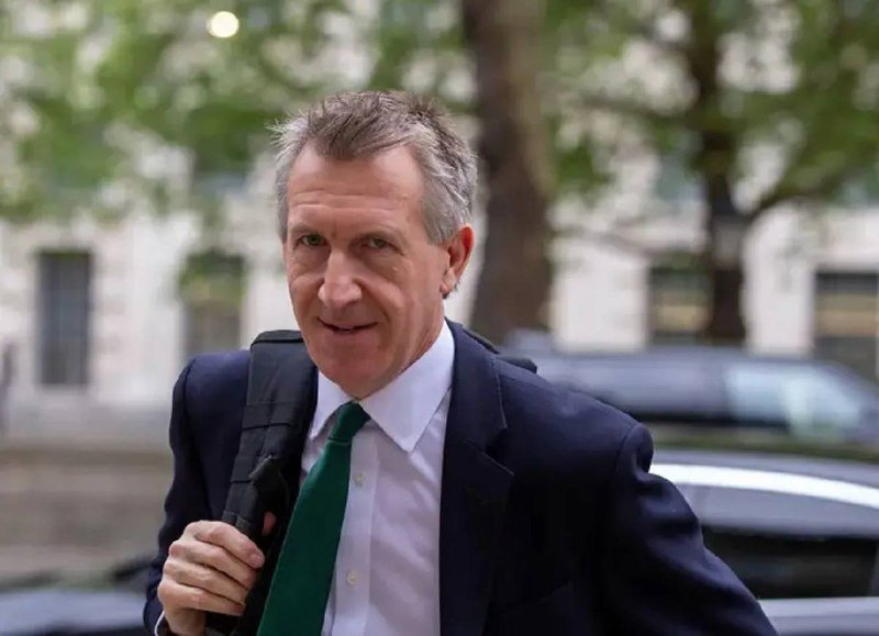 A gray-haired man in a dark suit and green tie walks outdoors carrying a black backpack strap over his shoulder.