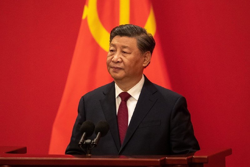 A man in a dark suit and red tie stands at a podium with microphones, positioned in front of a red flag with a yellow emblem.