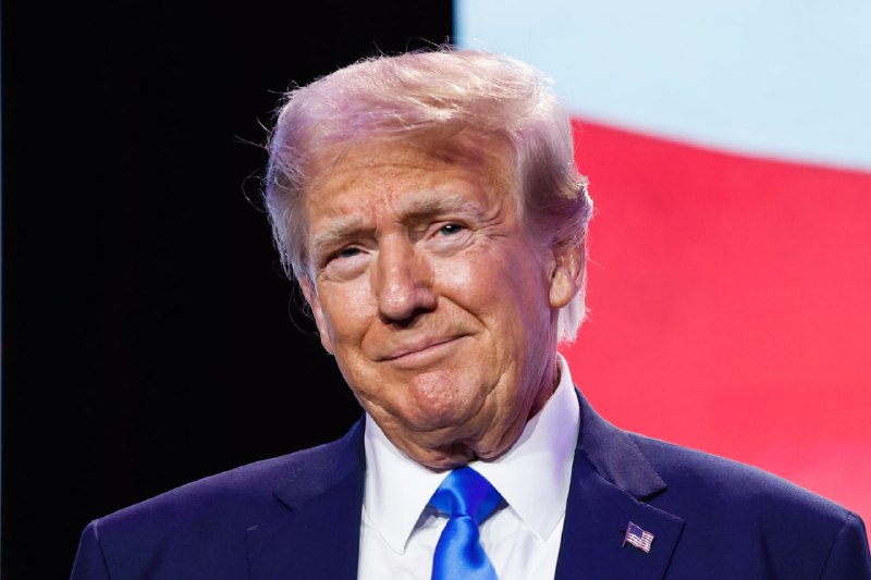 A blonde man in a navy suit and red tie gestures with both hands while speaking, with formally dressed people standing behind him.
