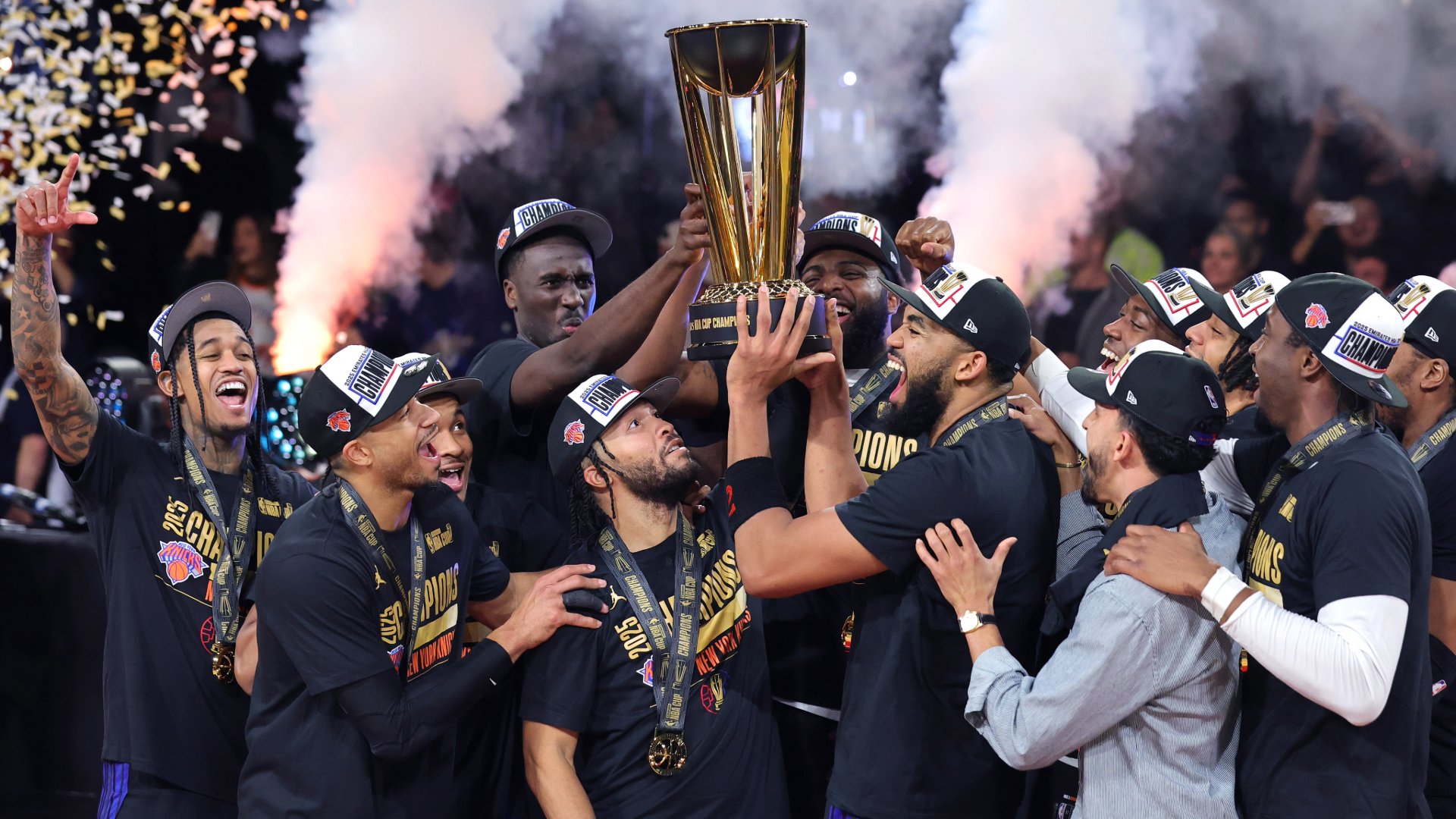 Basketball players in black championship shirts and hats celebrate by hoisting a gold NBA Cup trophy amid smoke and confetti.